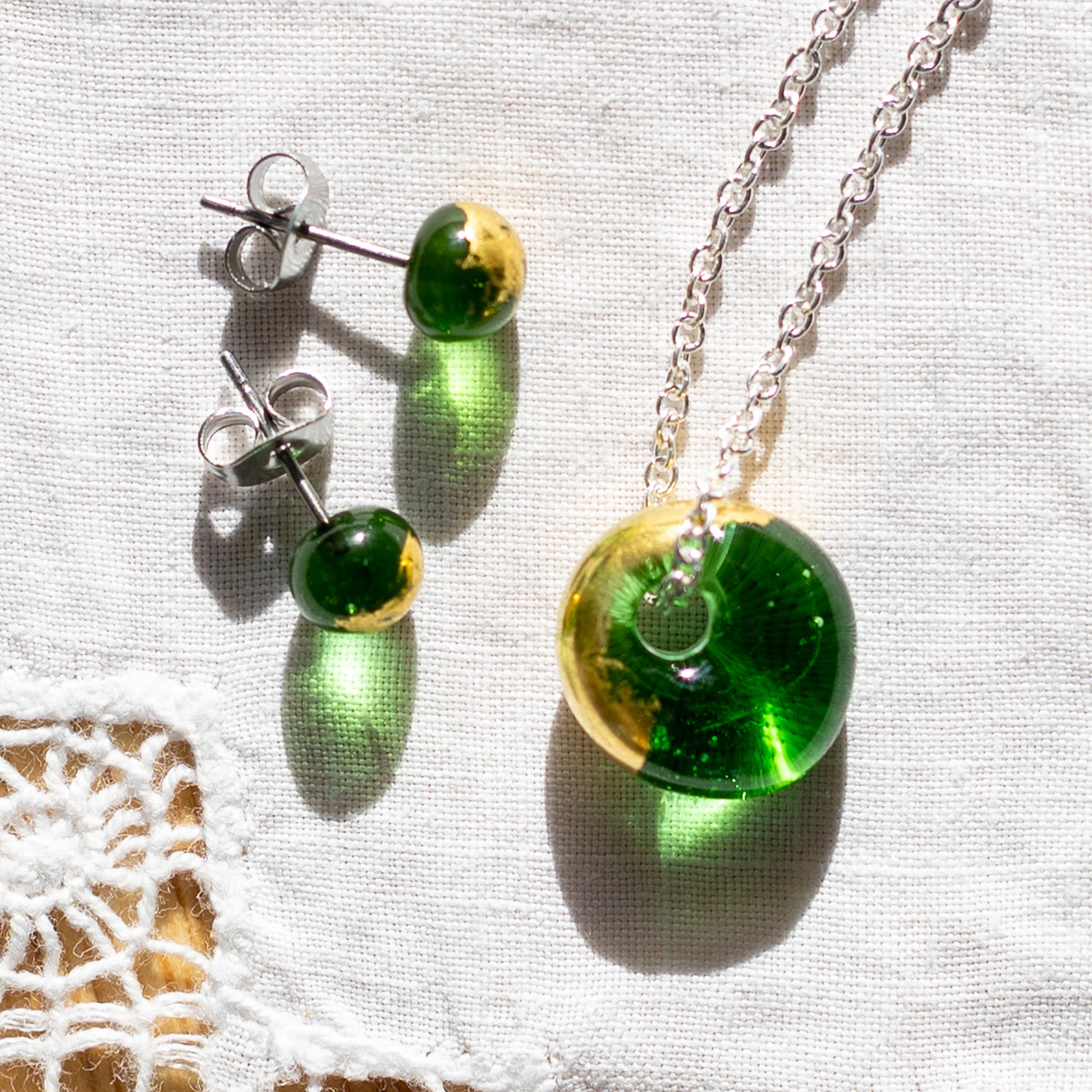 A jewellery set featuring a silver chain necklace and matching stud earrings with round green and gold glass pendant, displayed on a textured white fabric background.