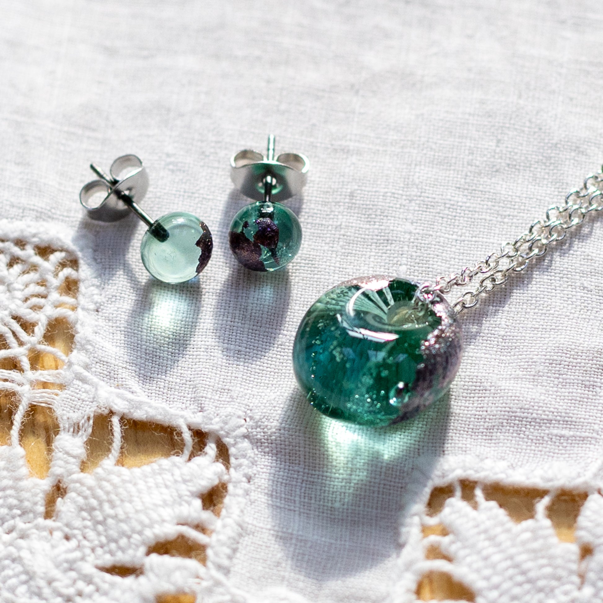 A jewellery set featuring a round green glass pendant necklace and matching round glass stud earrings with silver posts, displayed on a white textured fabric with a lace pattern.