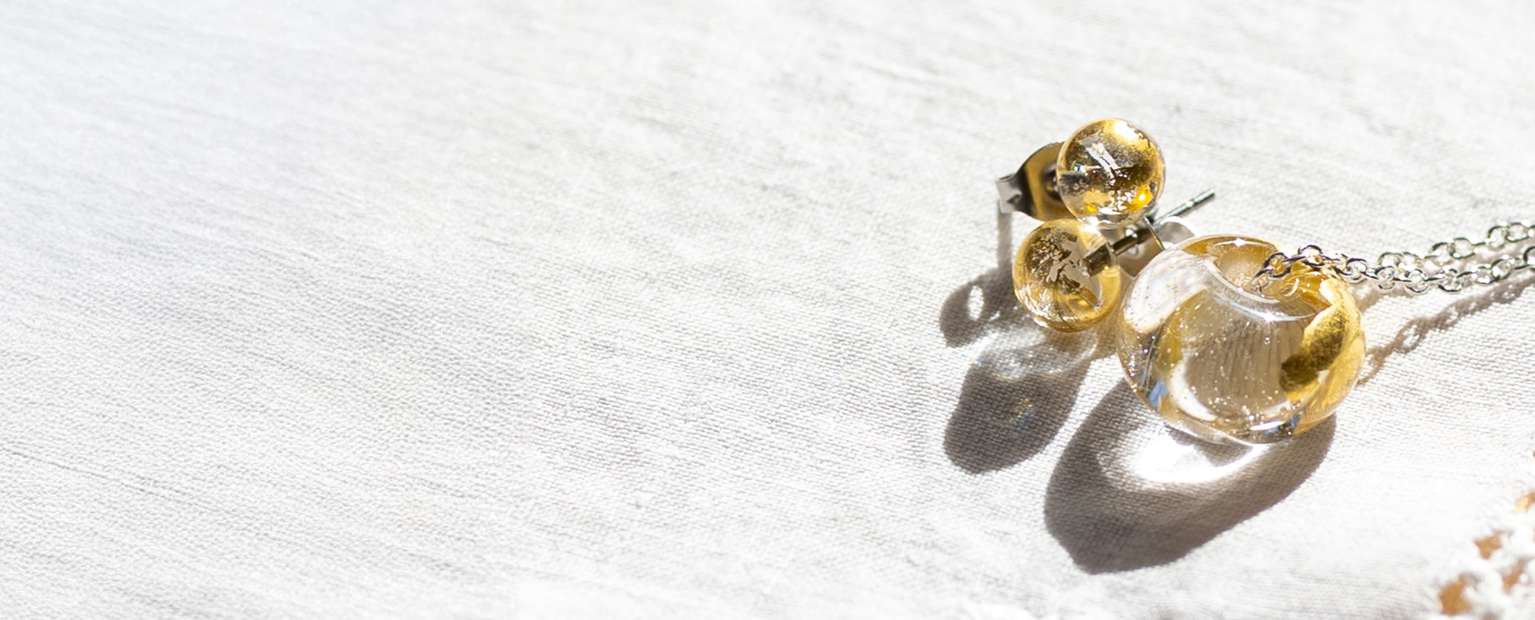 Recycled whisky bottle clear glass and gold pendant and matching clear glass stud earrings on a silver chain, displayed on a textured white fabric under bright sunlight.