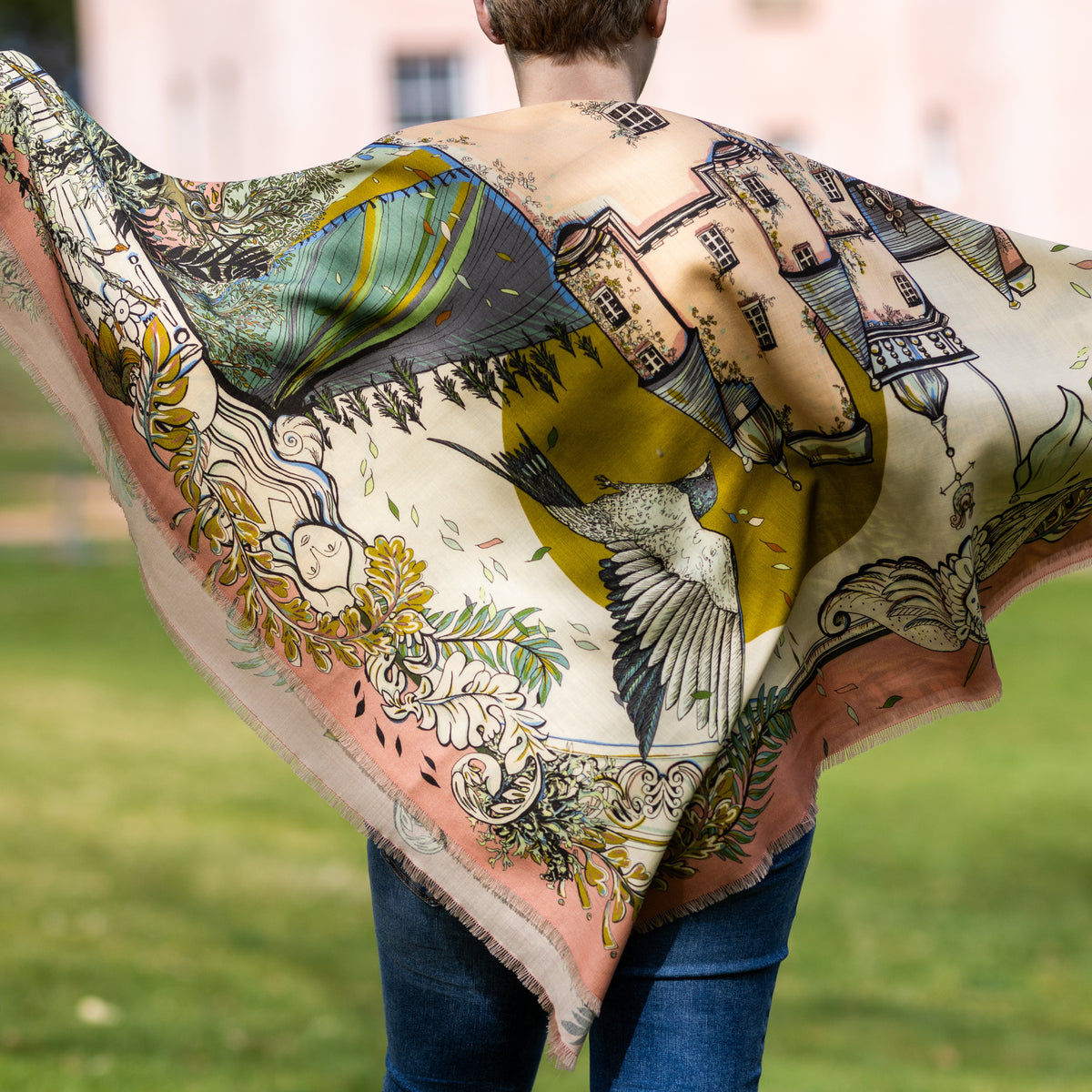 A person outdoors wearing a large printed scarf spread wide behind them. The fabric has an ornate illustrated pattern with Craigievar Castle in the centre, birds, foliage, and floral motifs in beige, pink, green, blue, and yellow tones.