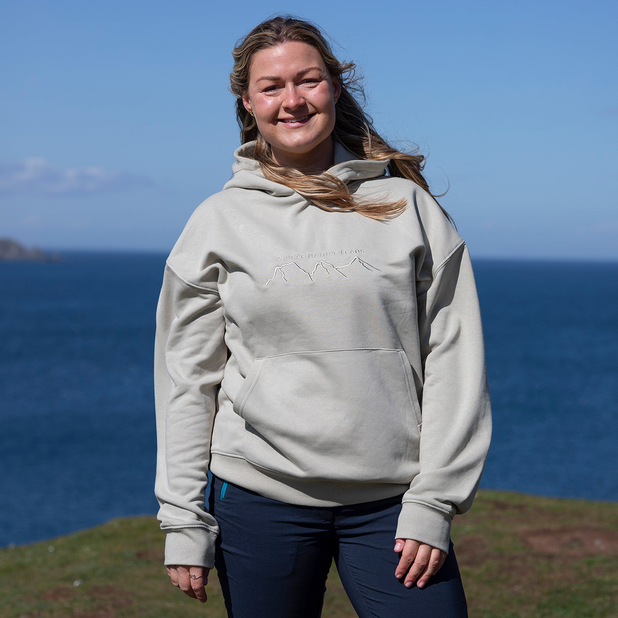 Lady standing on a cliff top with a blue sea backdrop wearing a Keela 'Where Nature Leads' Hoodie