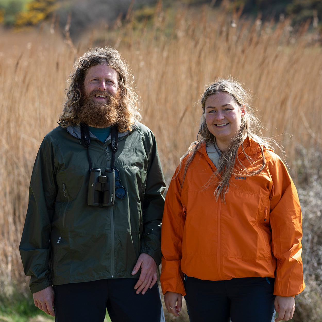 Two people standing in a field of tall grass, wearing Keela Outdoors jackets with one in green and the other an orange.