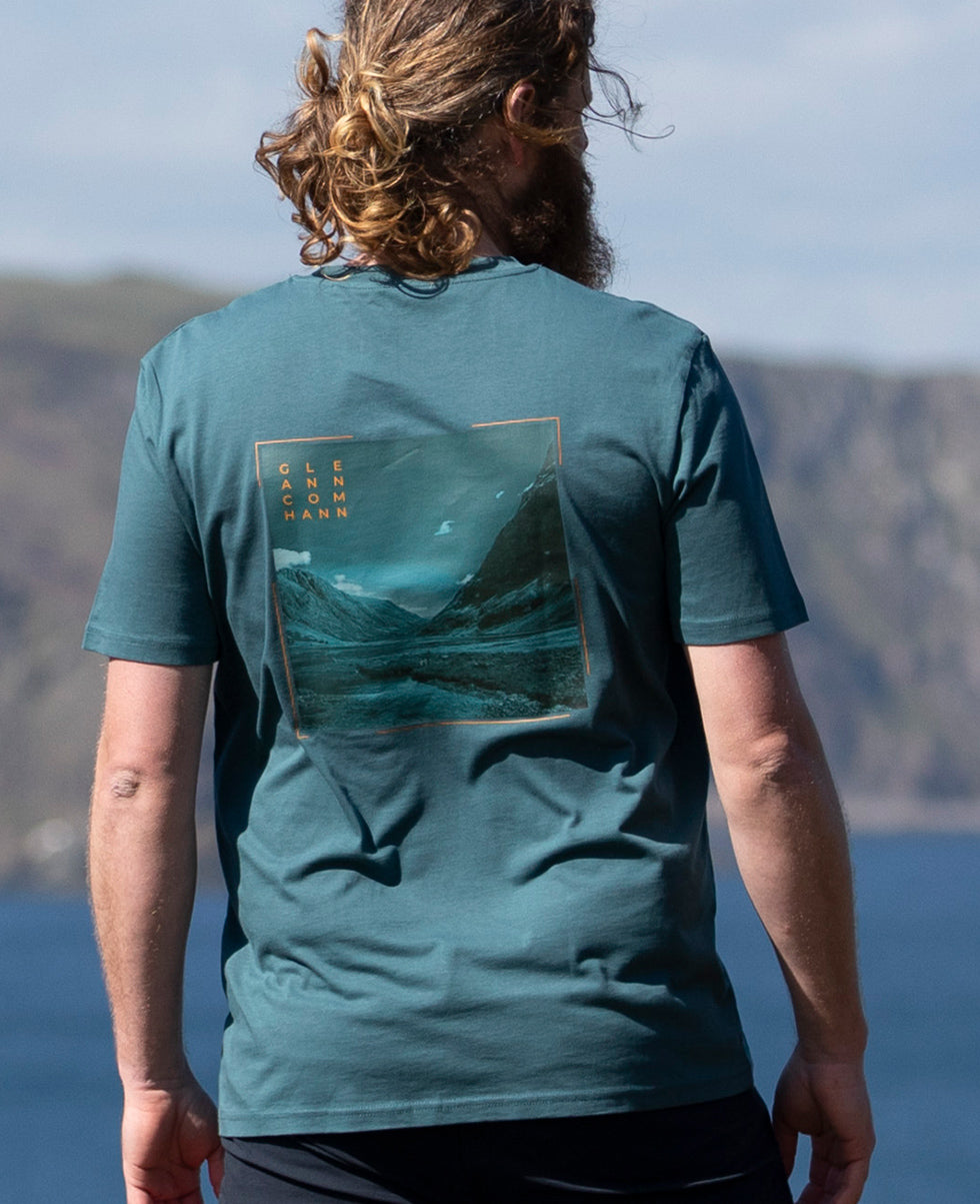 Person wearing a teal Keela Men's Glencoe T-Shirt with a mountain graphic and text, standing outdoors.