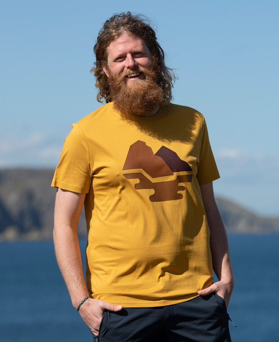 Man wearing a mustard Keela Men's St Kilda T-Shirt with a brown logo standing by a body of water.