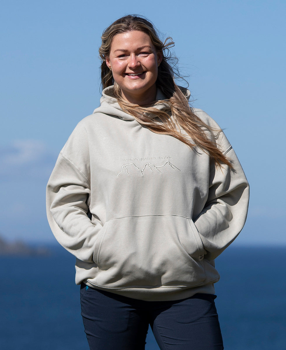 Person wearing a Keela 'Where Nature Leads' beige hoodie with a logo, standing against a blue sky and ocean background