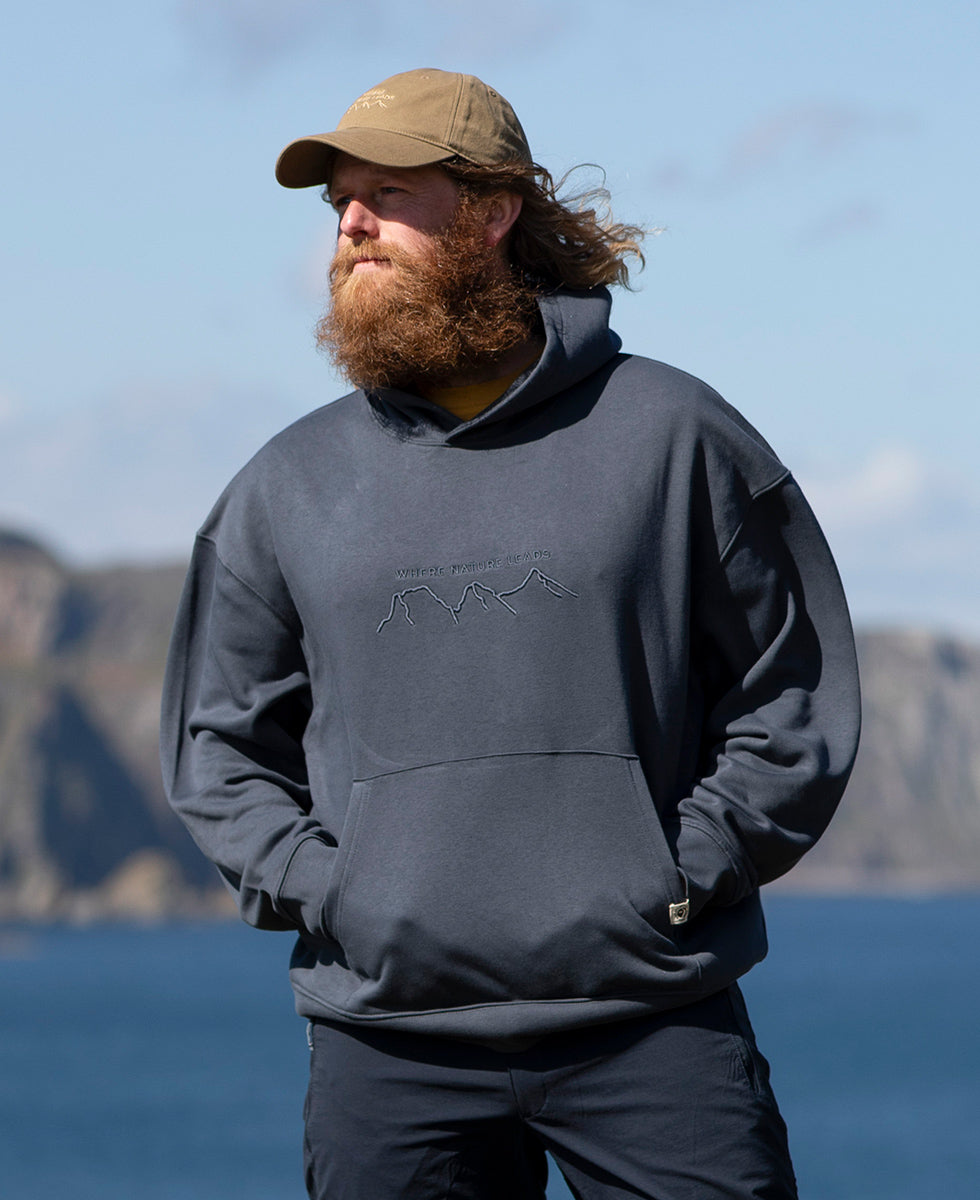 Man wearing a gray Keela 'Where Nature Leads' hoodie with a logo, standing outdoors with mountains and water in the background
