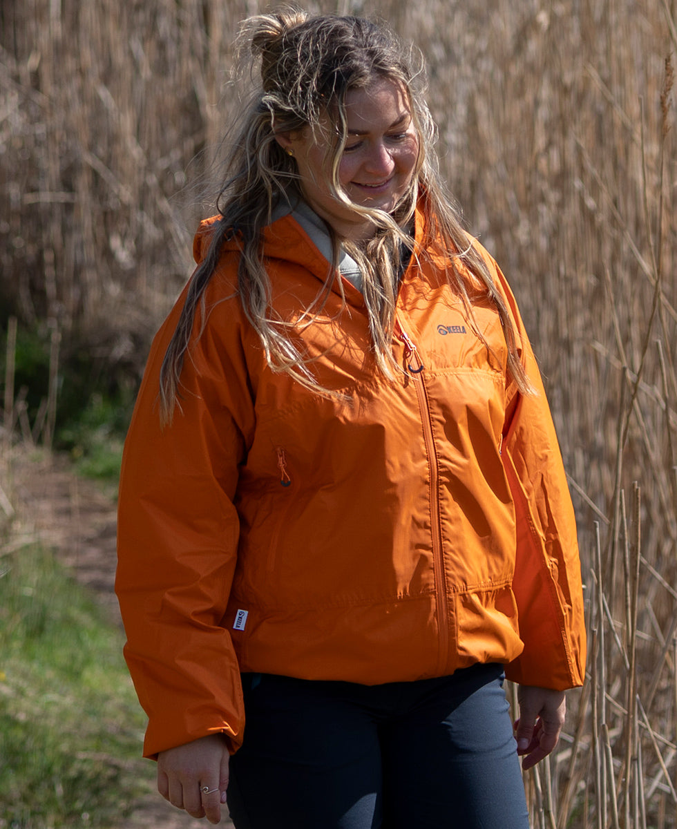 Woman wearing an orange Keela 'Where Nature Leads' packaway jacket standing in a natural setting with tall grass.