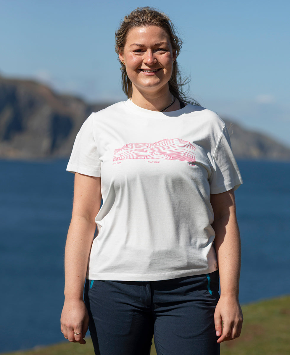 Woman wearing a white Keela Women's Ben Lomond T-Shirt with a pink design, standing in front of a scenic background with mountains and water.