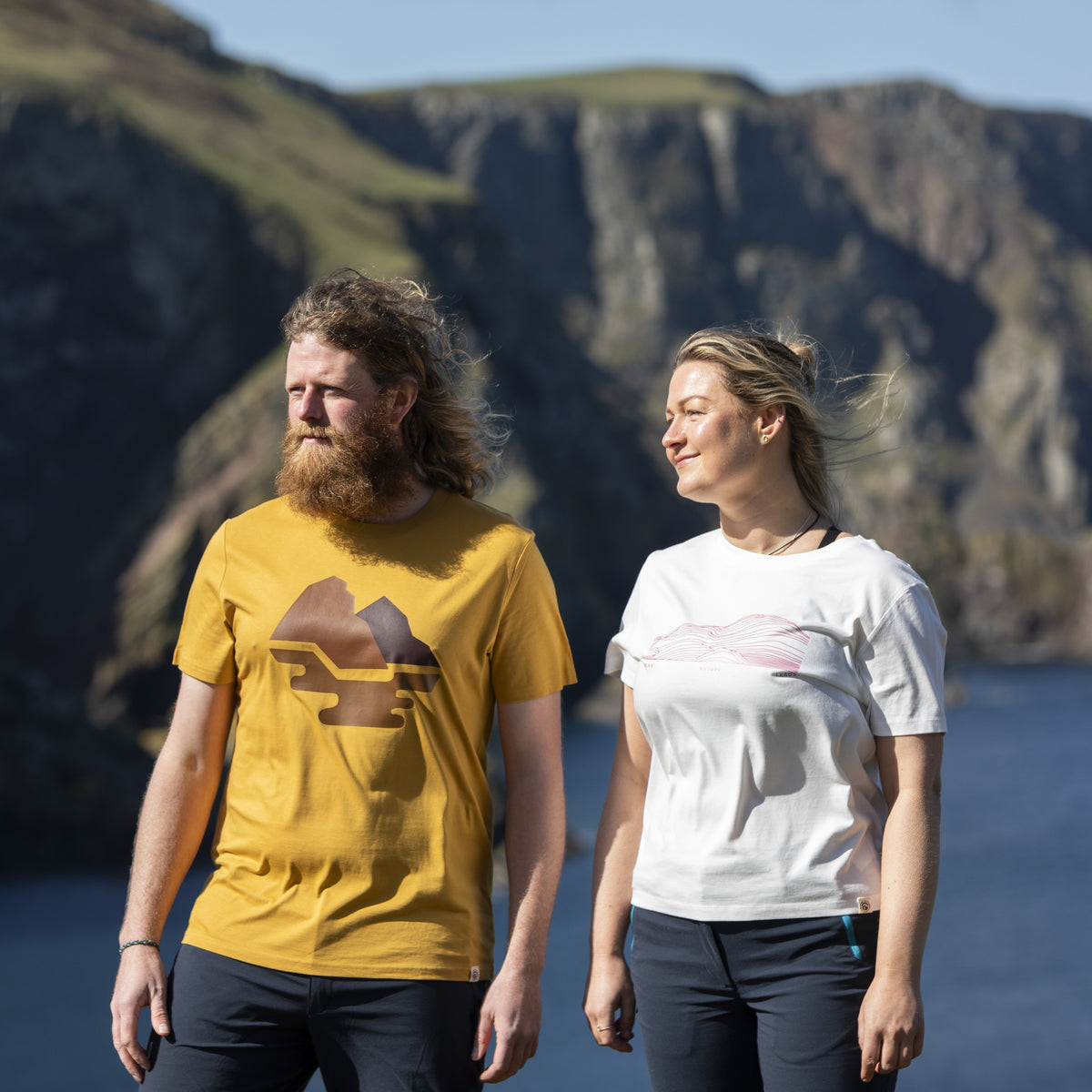 Two people walking outdoors in front of a rocky cliffside and water, wearing graphic T-shirts. The person on the left wears a mustard yellow shirt with a brown mountain graphic, and the person on the right wears a white shirt with a pale pink graphic.