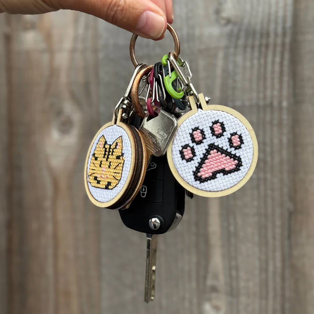 Keychain with cross-stitched charms featuring a cat and paw print design, held against a wooden background.