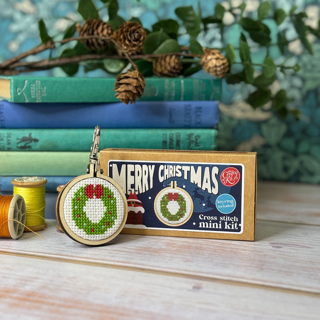 Cross stitch mini kit with Christmas wreath design, packaging, and embroidery hoop on a wooden surface with books and greenery in the background.