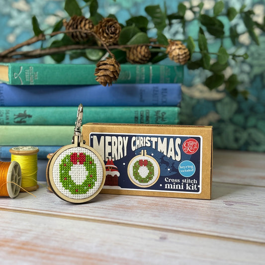 Cross stitch mini kit with Christmas wreath design, packaging, and embroidery hoop on a wooden surface with books and greenery in the background.