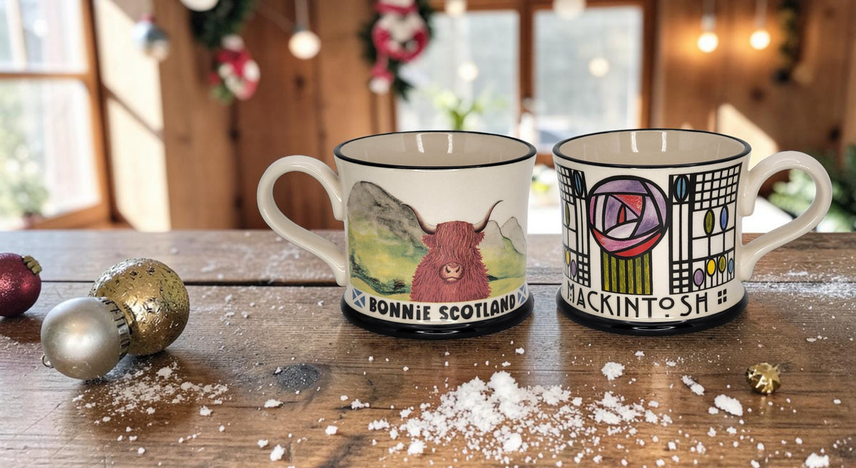 Two ceramic mugs with hand-painted Scottish illustrations, displayed on a rustic wooden table with festive holiday decorations in the background. One mug features a red highland cow on a green landscape with the text 'BONNIE SCOTLAND', and the other features a colorful abstract design with the text 'MACKINTOSH'.