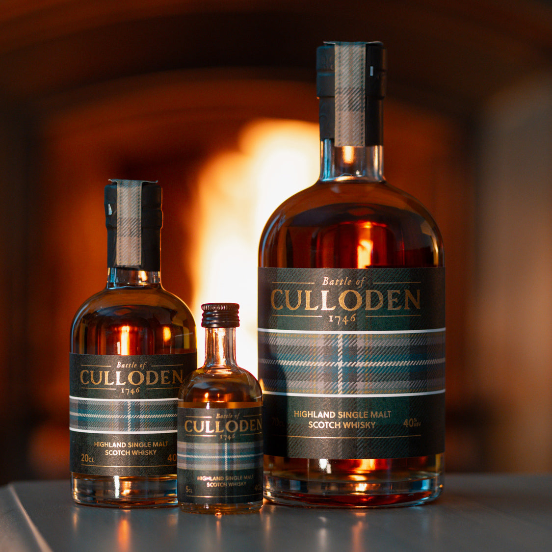 Three bottles of Culloden Highland Single Malt Scotch Whisky are arranged on a reflective surface against a warm, blurred background. The set includes a large bottle, a medium bottle, and a small bottle, all featuring the Spirit of Culloden Tartan.