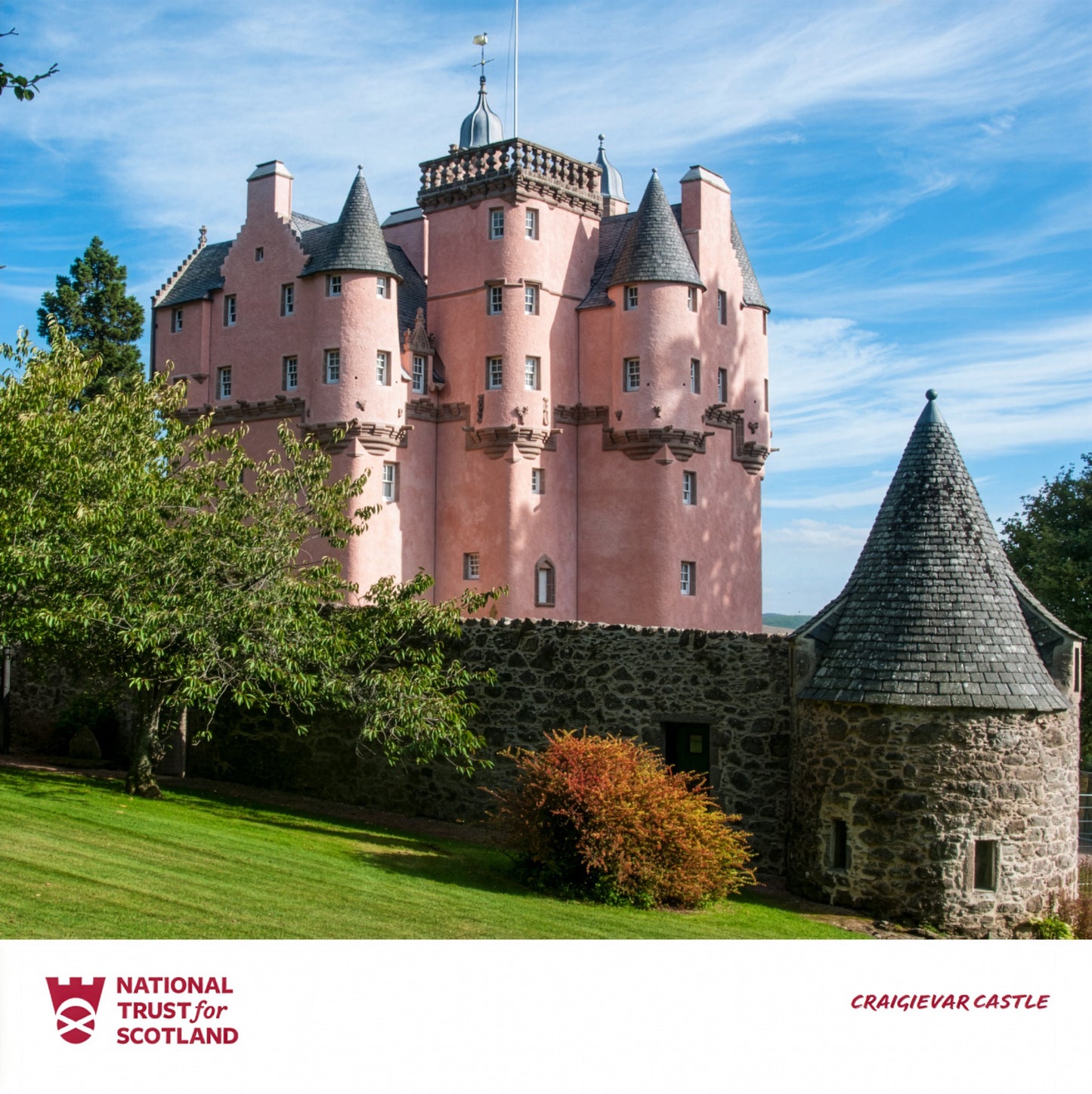 Craigievar 'Pink' castle with turrets and a stone wall, surrounded by greenery, under a blue sky.