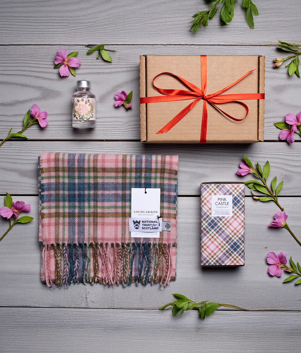 Gift set with a Pink Castle tartan scarf, box, and a miniature bottle of Pink Castle gin on a wooden surface with flowers.