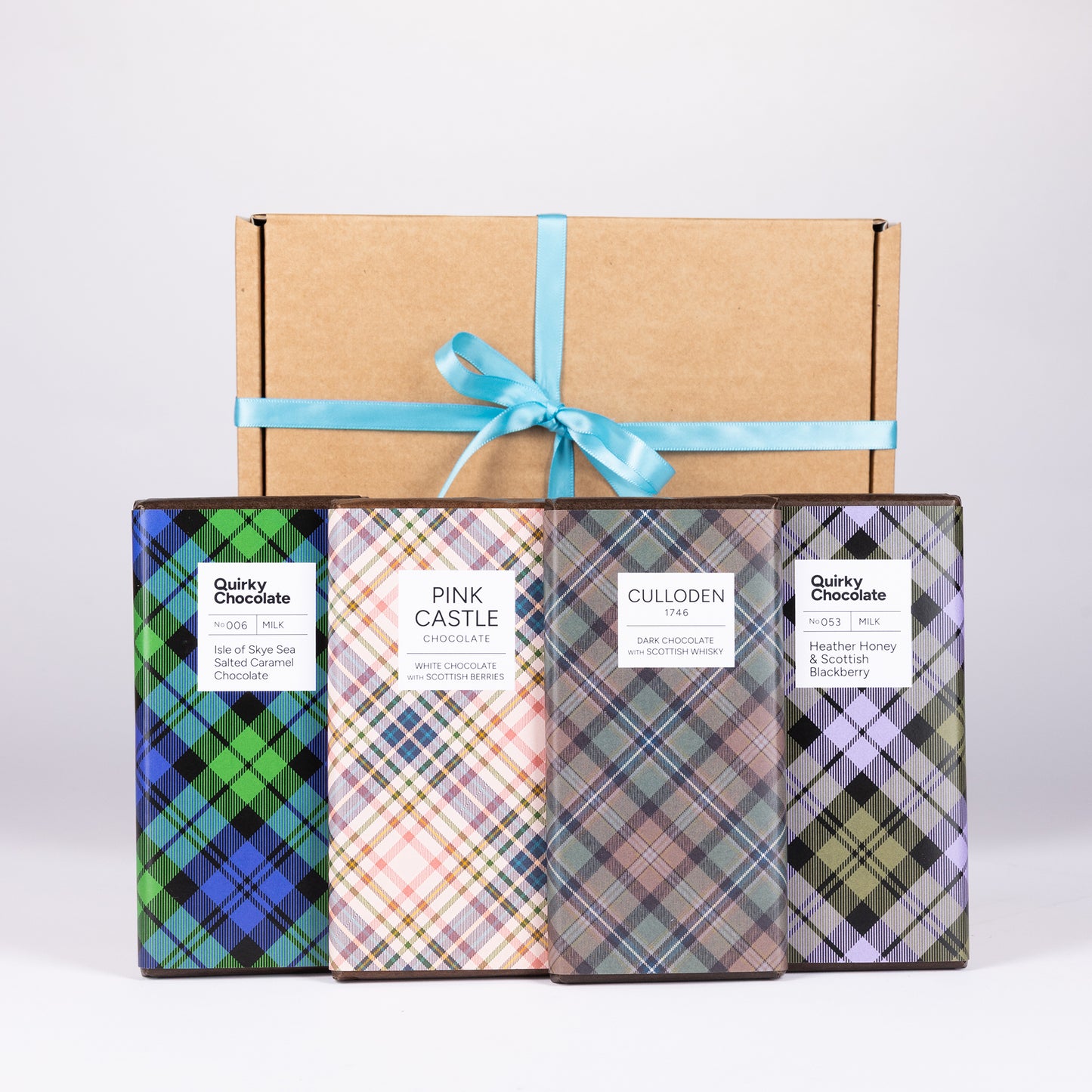 Four Quirky chocolate bars in individually wrapped plaid boxes are displayed in front of a plain brown cardboard gift box tied with a light teal ribbon.