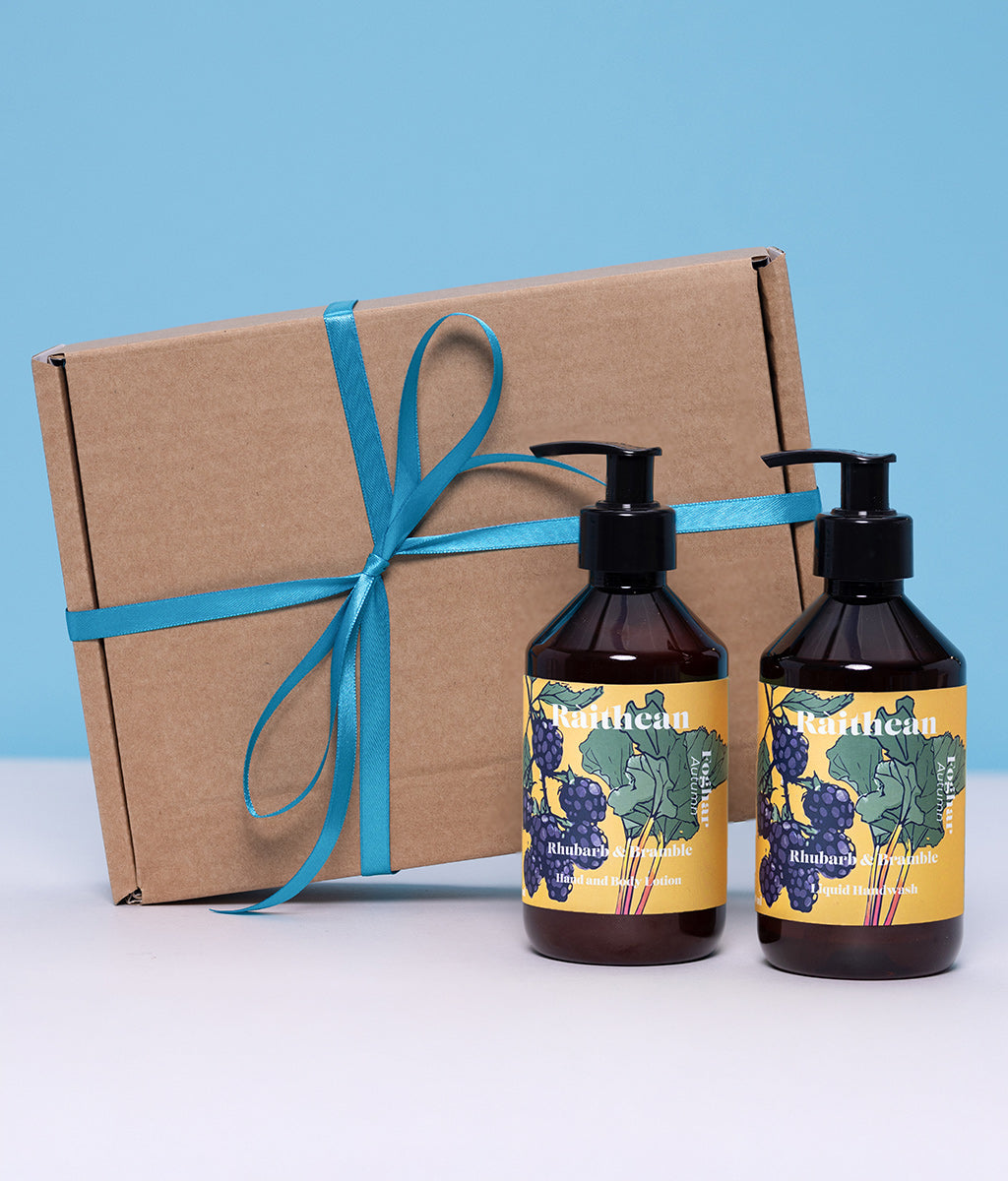 Two Raithean Autumn soap bottles with yellow labels next to a cardboard box tied with teal ribbon on a light blue background.