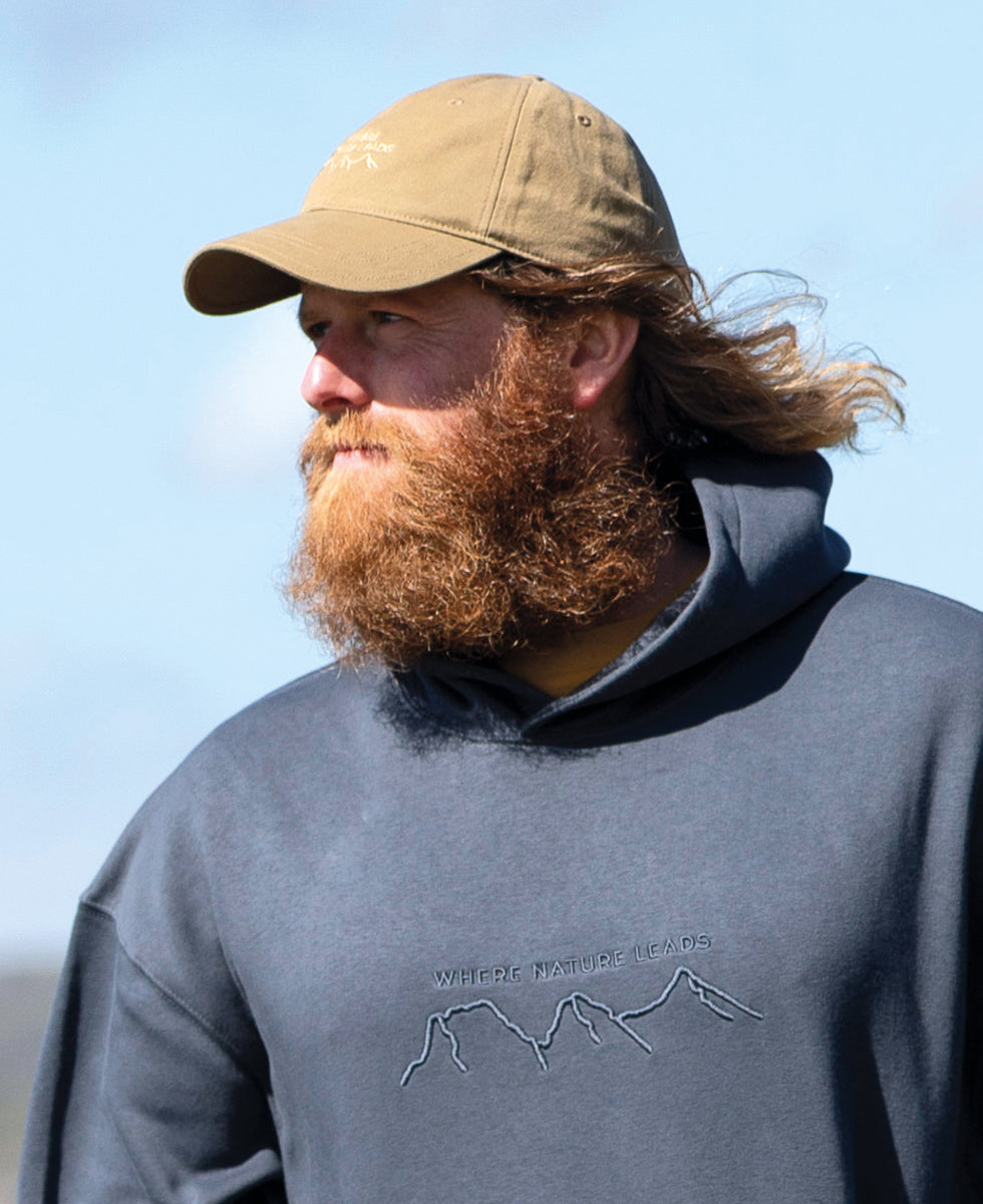 Man wearing a 'Where Nature Leads' baseball cap and gray hoodie with mountain design outdoors