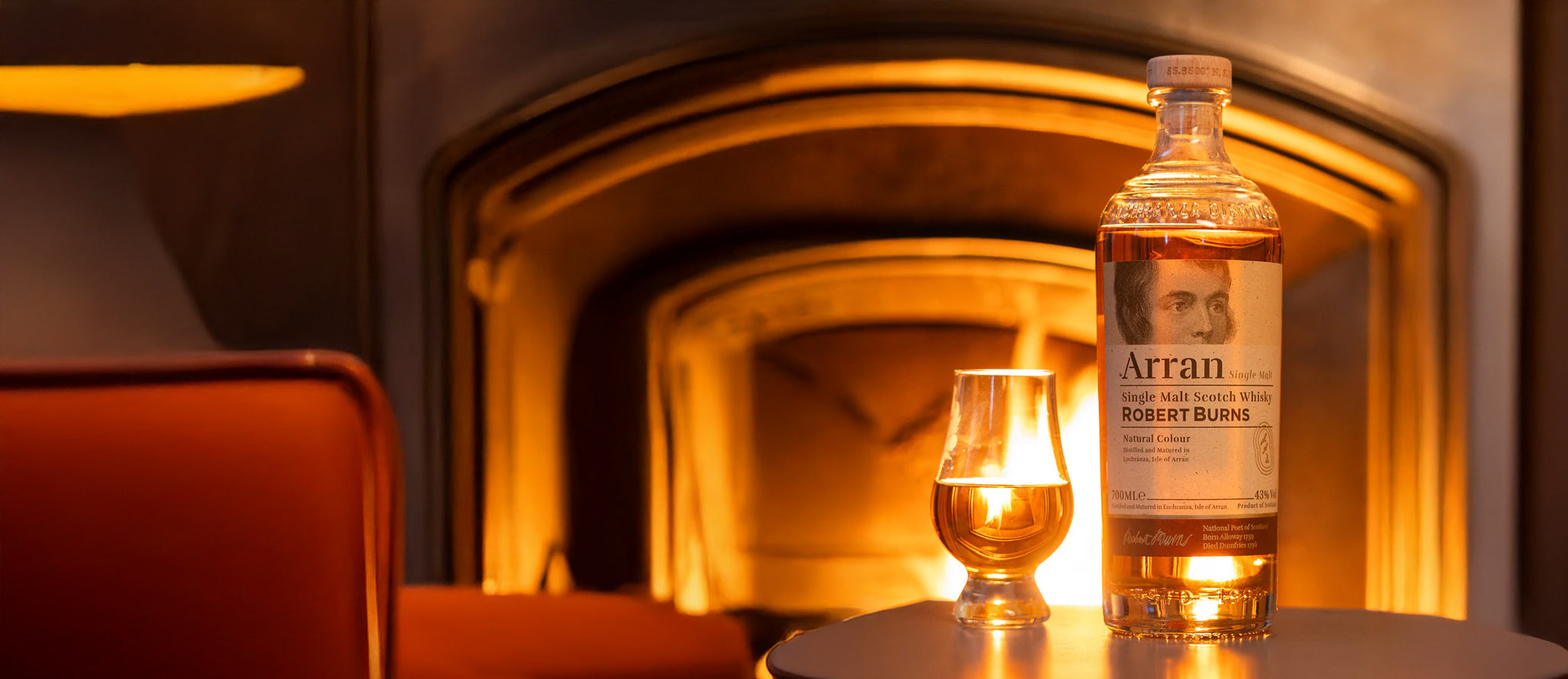 Robert Burns Arran Single Malt Whisky and glass sitting on table in front of a flaming fireplace.
