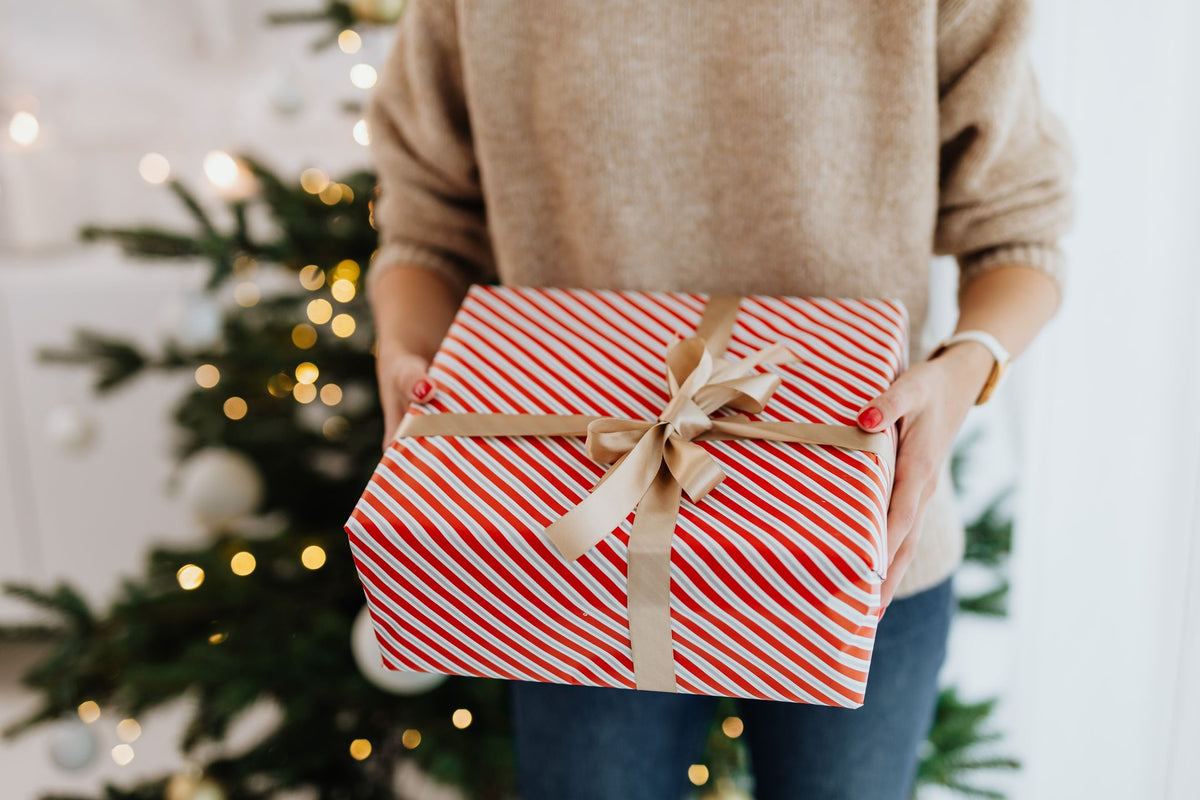 A person in a beige sweater holds a wrapped gift box with red and white diagonal stripes and a tan ribbon bow, with a blurred Christmas tree in the background.