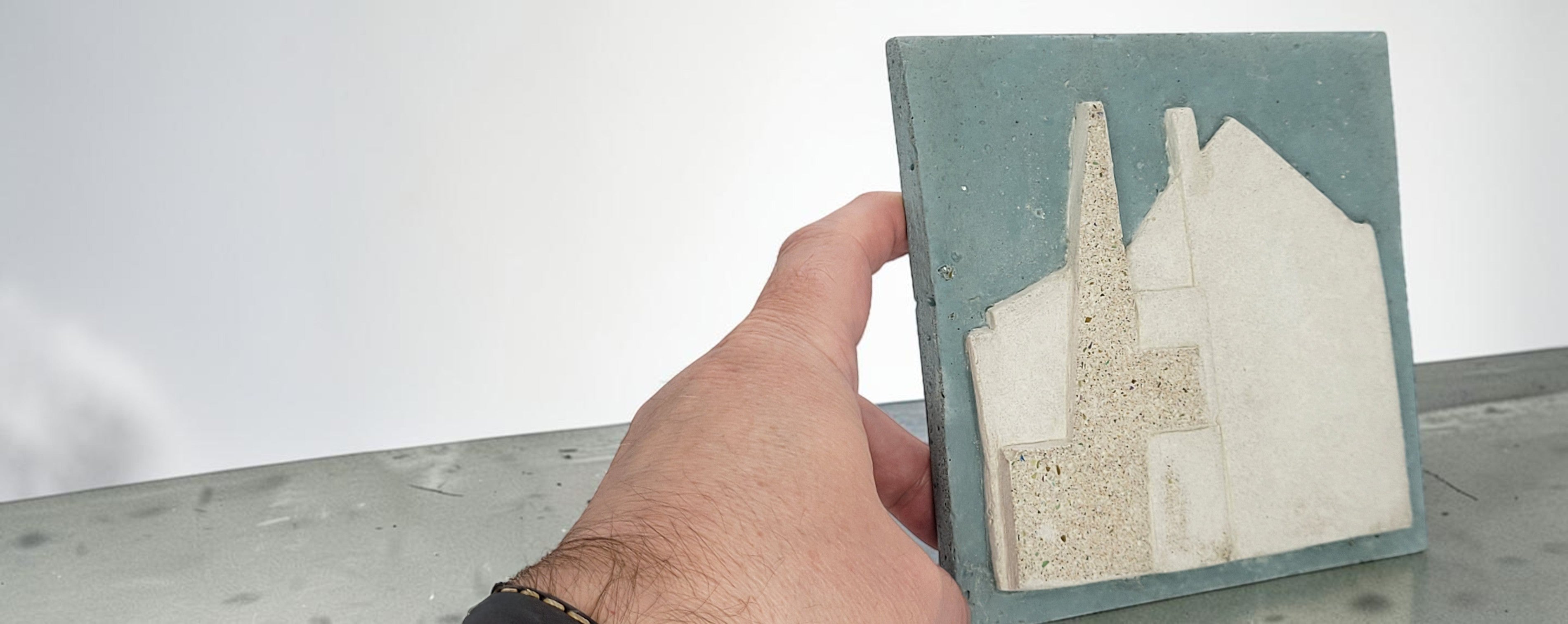 A hand holding a square ceramic tile featuring a textured, raised architectural design of the Mackintosh Hill House hanging wall tile in cream and beige tones against a muted teal-green background.